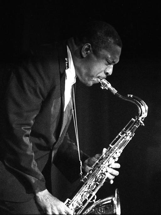 Historisches Schwarzweißfoto von John Coltrane, der auf einer Bühne steht und Saxophon spielt.