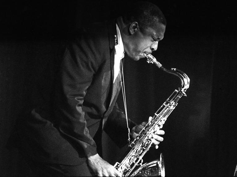 Historisches Schwarzweißfoto von John Coltrane, der auf einer Bühne steht und Saxophon spielt.