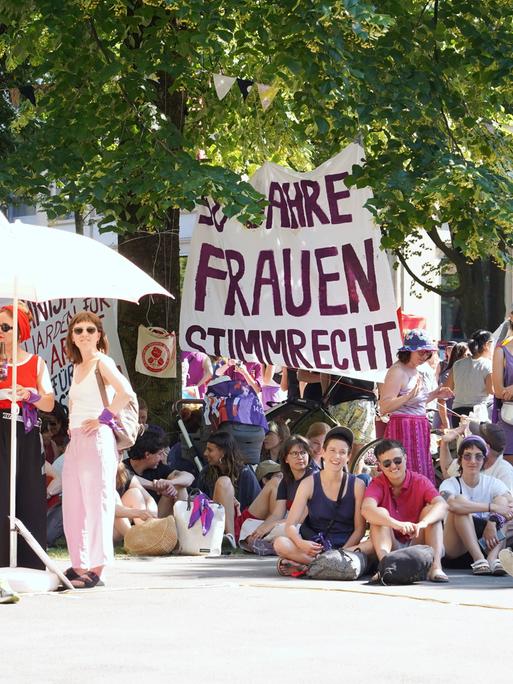 Frauen sitzen und stehen im Schatten von Bäumen und Sonnenschirmen, im Hintergrund ist ein Transparent mit der Aufschrift "50 Jahre Frauenwahlrecht" zu sehen.