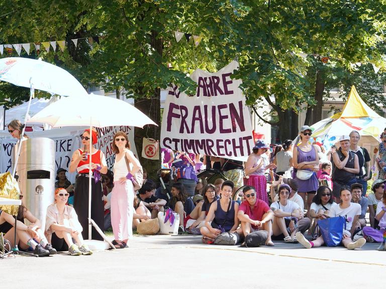 Frauen sitzen und stehen im Schatten von Bäumen und Sonnenschirmen, im Hintergrund ist ein Transparent mit der Aufschrift "50 Jahre Frauenwahlrecht" zu sehen.