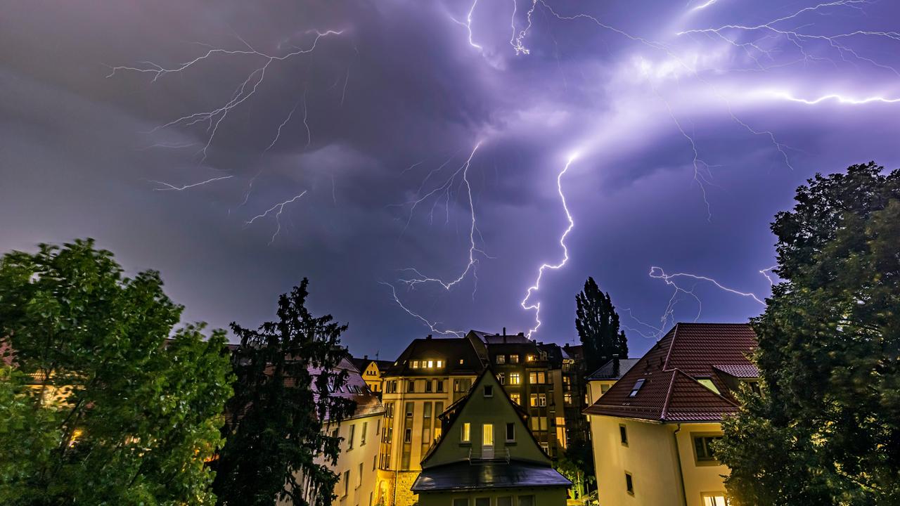 Ein Gewitter über einer Stadt. Blitze zuucken aus dem Himmel. Es ist Nacht. In vielen Fenstern der Häuser brennt Licht. 