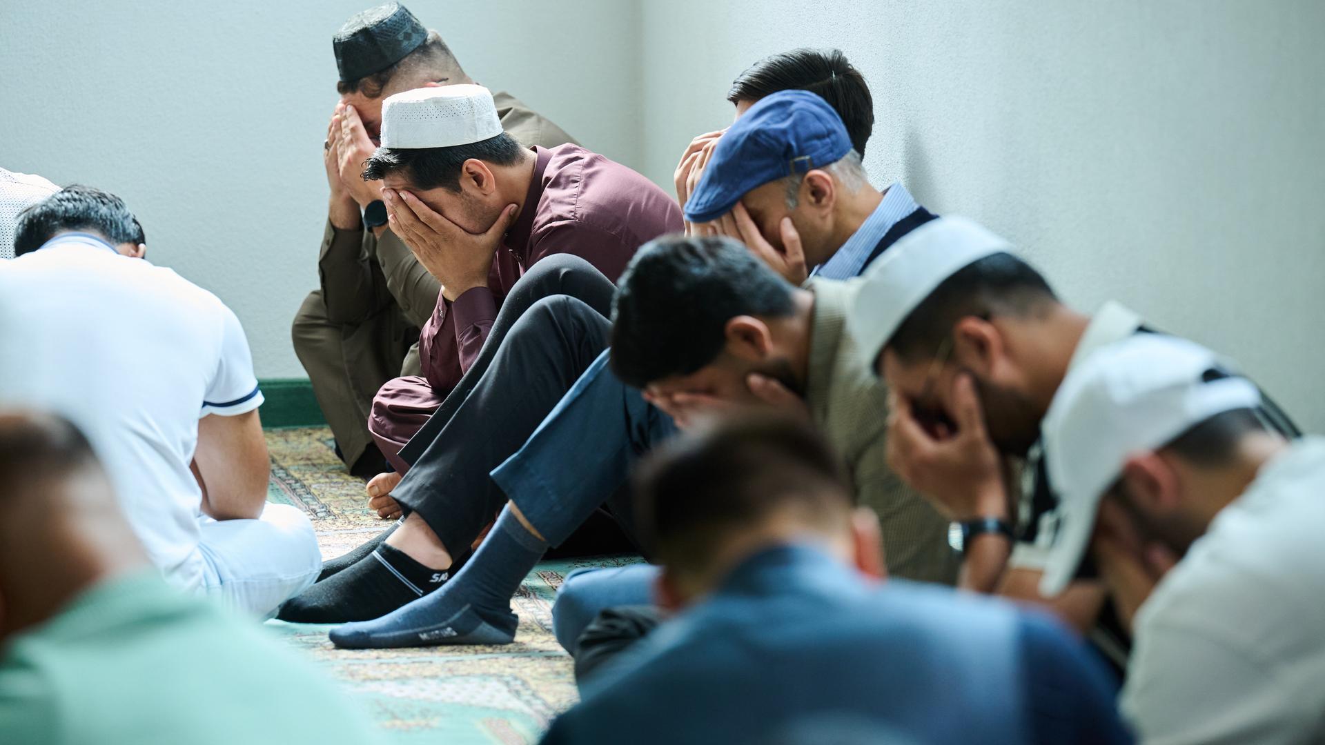 Muslimische Männer beten in der Moschee. Mehrere Männer halten zum Gebet das Gesicht verdeckt und sitzen auf dem Boden. 