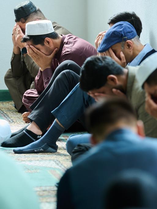Muslimische Männer beten in der Moschee. Mehrere Männer halten zum Gebet das Gesicht verdeckt und sitzen auf dem Boden. 