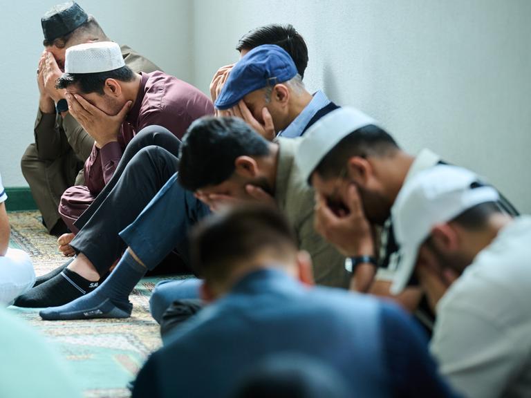 Muslimische Männer beten in der Moschee. Mehrere Männer halten zum Gebet das Gesicht verdeckt und sitzen auf dem Boden. 