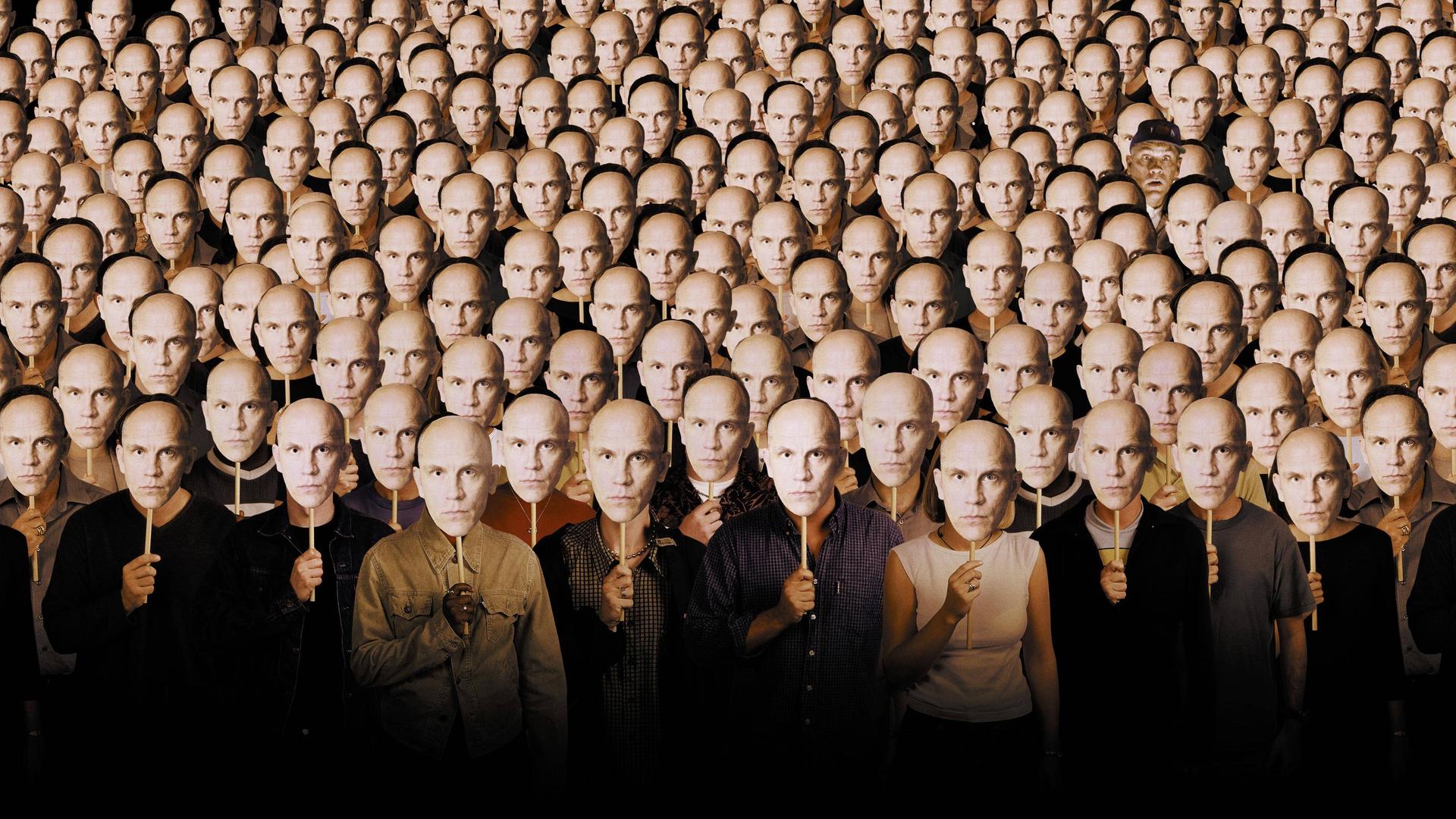 Im Filmbild aus "Being John Malkovich" stehen viele Menschen nebeneinander und halten sich Pappmasken vor die eigenen Gesichter, die alle das Gesicht von John Malkovich zeigen.