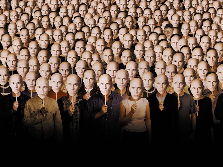 Im Filmbild aus "Being John Malkovich" stehen viele Menschen nebeneinander und halten sich Pappmasken vor die eigenen Gesichter, die alle das Gesicht von John Malkovich zeigen.