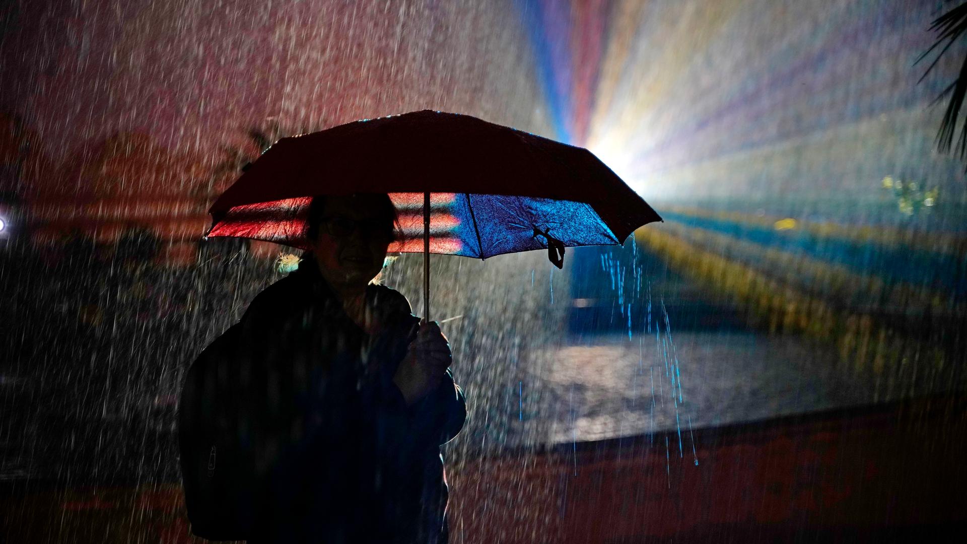 Ein Mensch steht in der Nacht mit einem Regenschirm im Gegenlicht. Das Licht hat verschiedene Farben. Dazu regnet es.