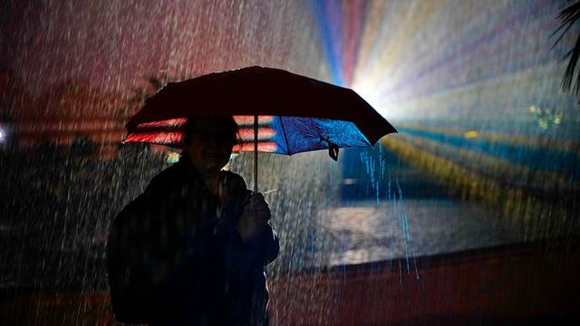 Ein Mensch steht in der Nacht mit einem Regenschirm im Gegenlicht. Das Licht hat verschiedene Farben. Dazu regnet es.