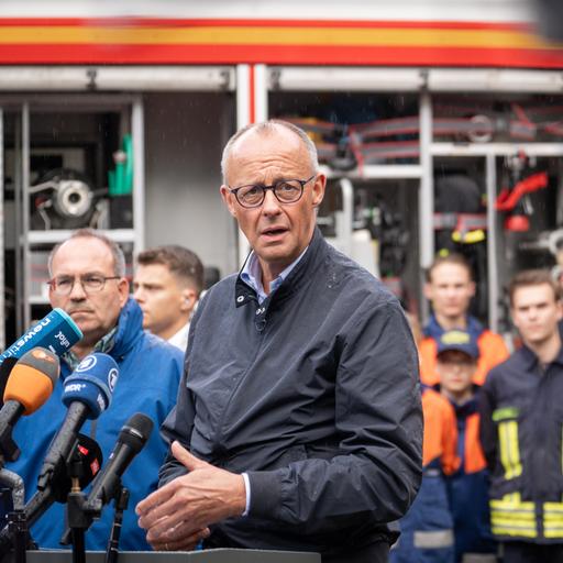 Merz spricht in mehrere Mikrofone. Hinter ihm sind der zuständige Landrat und mehrere Einsatzkräfte zu erkennen.