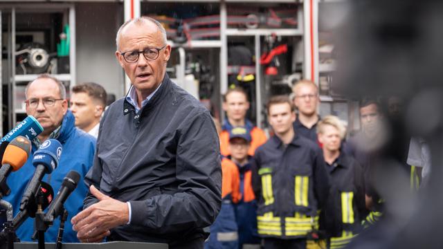 Merz spricht in mehrere Mikrofone. Hinter ihm sind der zuständige Landrat und mehrere Einsatzkräfte zu erkennen.