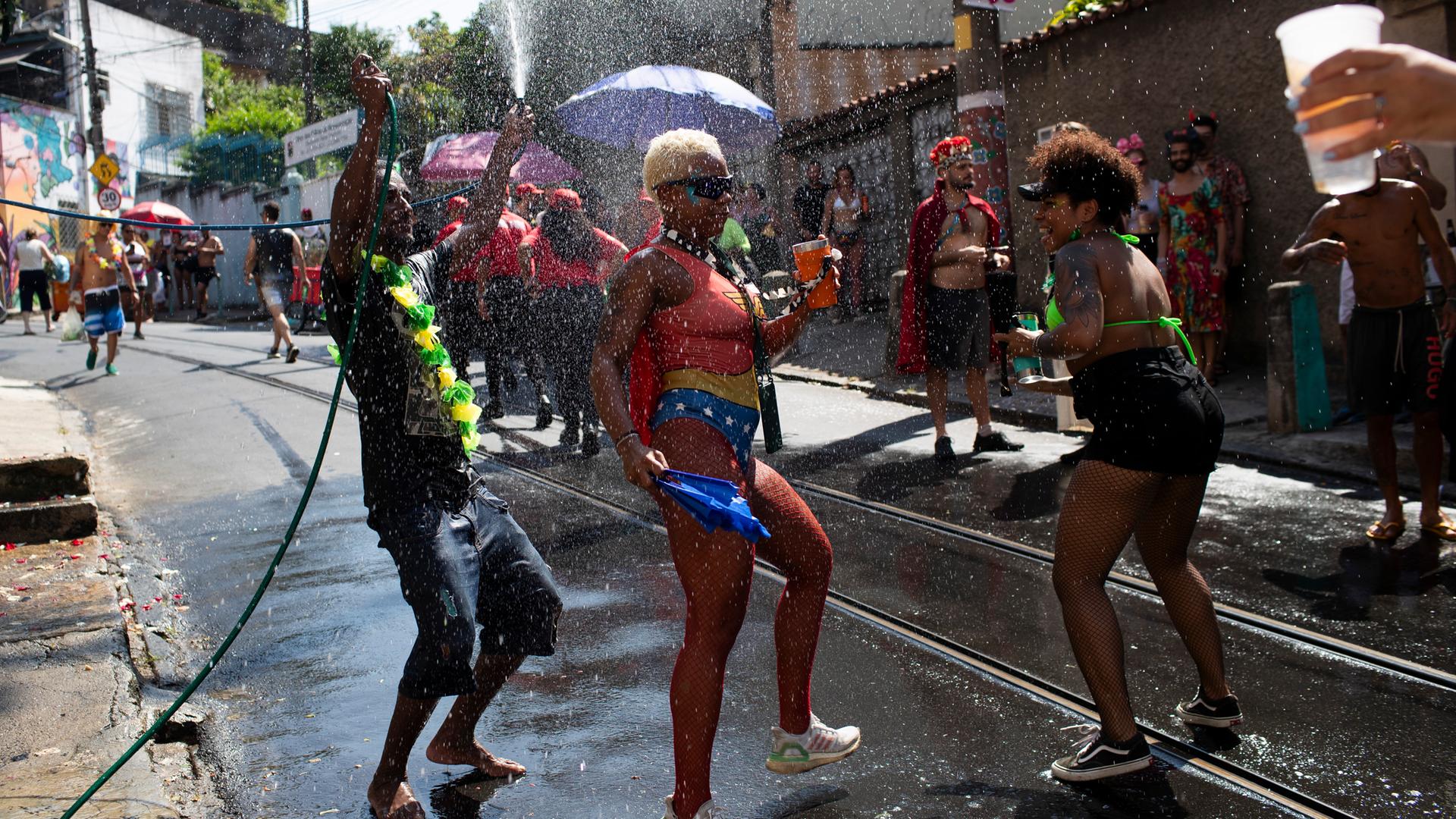 Drei kostümierte Menschen stehen auf einer Straße in Rio de Janeiro und feiern Karneval. Wegen der großen Hitze werden die Menschen zur Abkühlung  mit Wasser bespritzt. 