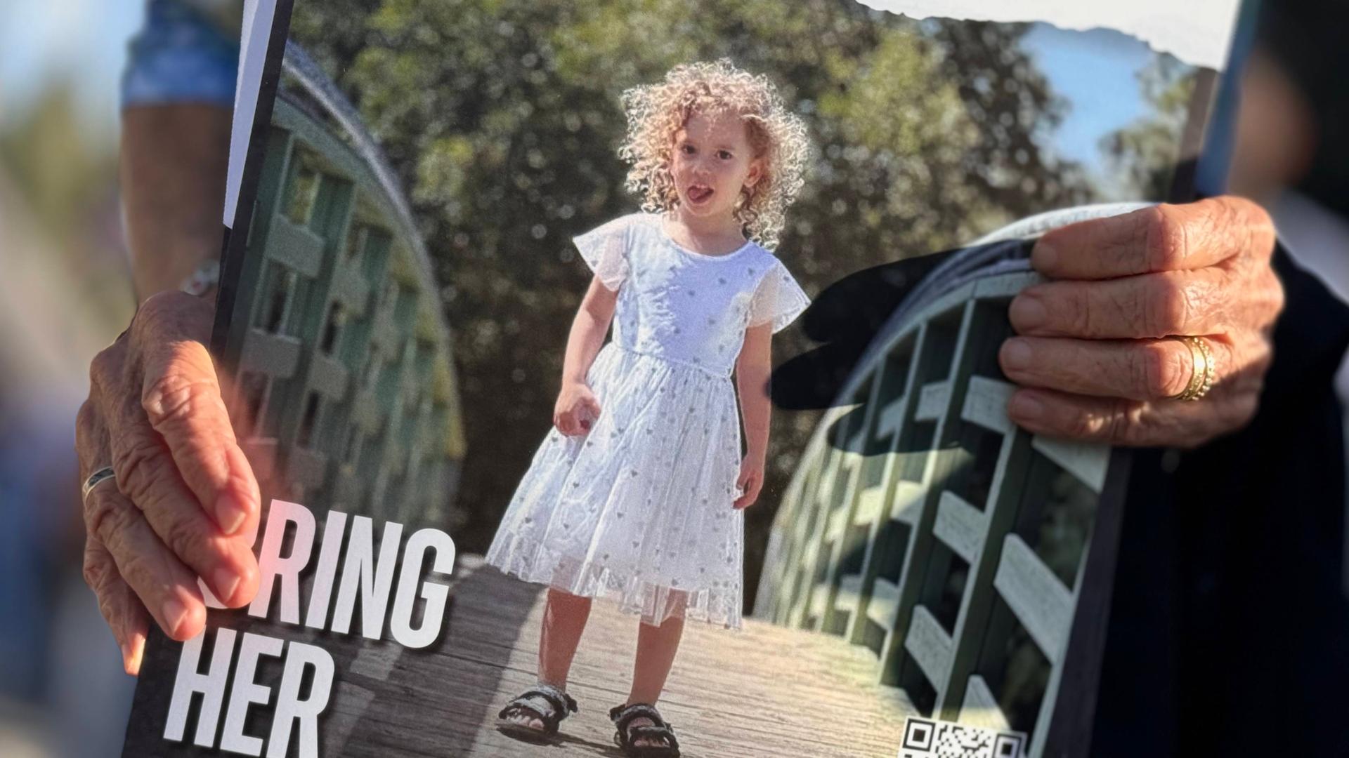Auf einem Plakat steht: "Bring Her Home Now!" Ein kleines Mädchen im Kleid ist darauf zu sehen, gehalten von alten Händen. 