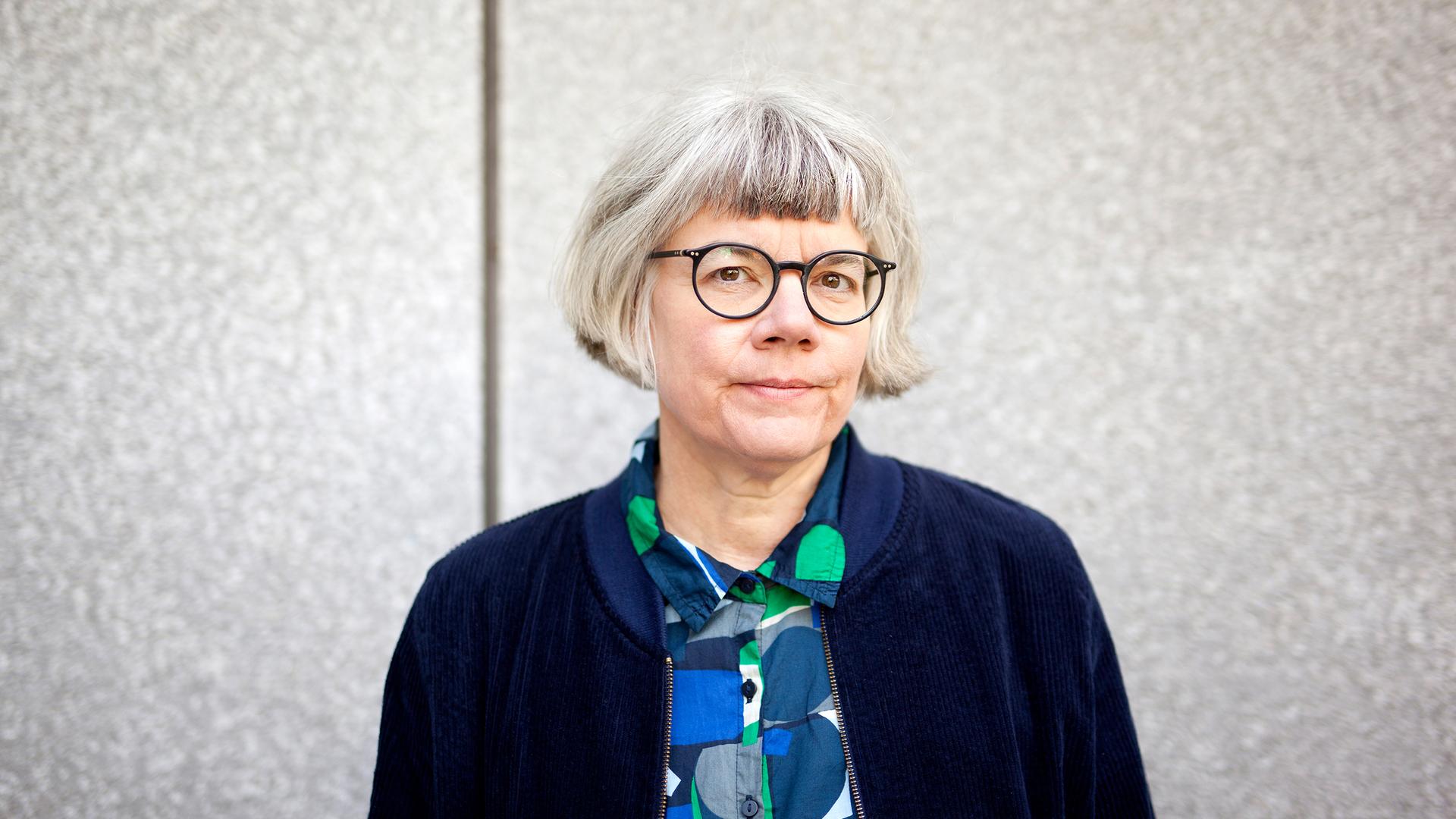 Eine Frau mit grauem Haar und Brille blickt ernst in die Kamera. Sie steht vor einem grauen Hintergrund und trägt eine dunkelblaue Jacke über einem blau-grünen Hemd. Eine Frau mit grauem Haar und Brille blickt ernst in die Kamera. Sie steht vor einem grauen Hintergrund und trägt eine dunkelblaue Jacke über einem blau-grünen Hemd.