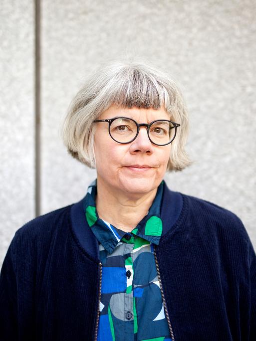 Eine Frau mit grauem Haar und Brille blickt ernst in die Kamera. Sie steht vor einem grauen Hintergrund und trägt eine dunkelblaue Jacke über einem blau-grünen Hemd. Eine Frau mit grauem Haar und Brille blickt ernst in die Kamera. Sie steht vor einem grauen Hintergrund und trägt eine dunkelblaue Jacke über einem blau-grünen Hemd.