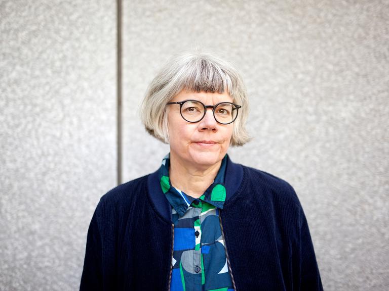 Eine Frau mit grauem Haar und Brille blickt ernst in die Kamera. Sie steht vor einem grauen Hintergrund und trägt eine dunkelblaue Jacke über einem blau-grünen Hemd. Eine Frau mit grauem Haar und Brille blickt ernst in die Kamera. Sie steht vor einem grauen Hintergrund und trägt eine dunkelblaue Jacke über einem blau-grünen Hemd.