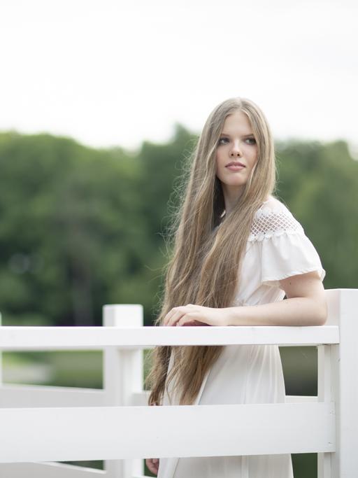 Eine junge Frau mit sehr langen blonden Haaren lehnt an eine weiße Brüstung. Sie trägt ein weißes Sommerkleid und blickt versonnen. Hinter ihr sind verschwommen Bäume zu erkennen.