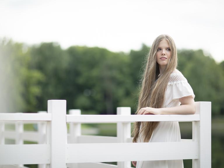 Eine junge Frau mit sehr langen blonden Haaren lehnt an eine weiße Brüstung. Sie trägt ein weißes Sommerkleid und blickt versonnen. Hinter ihr sind verschwommen Bäume zu erkennen.