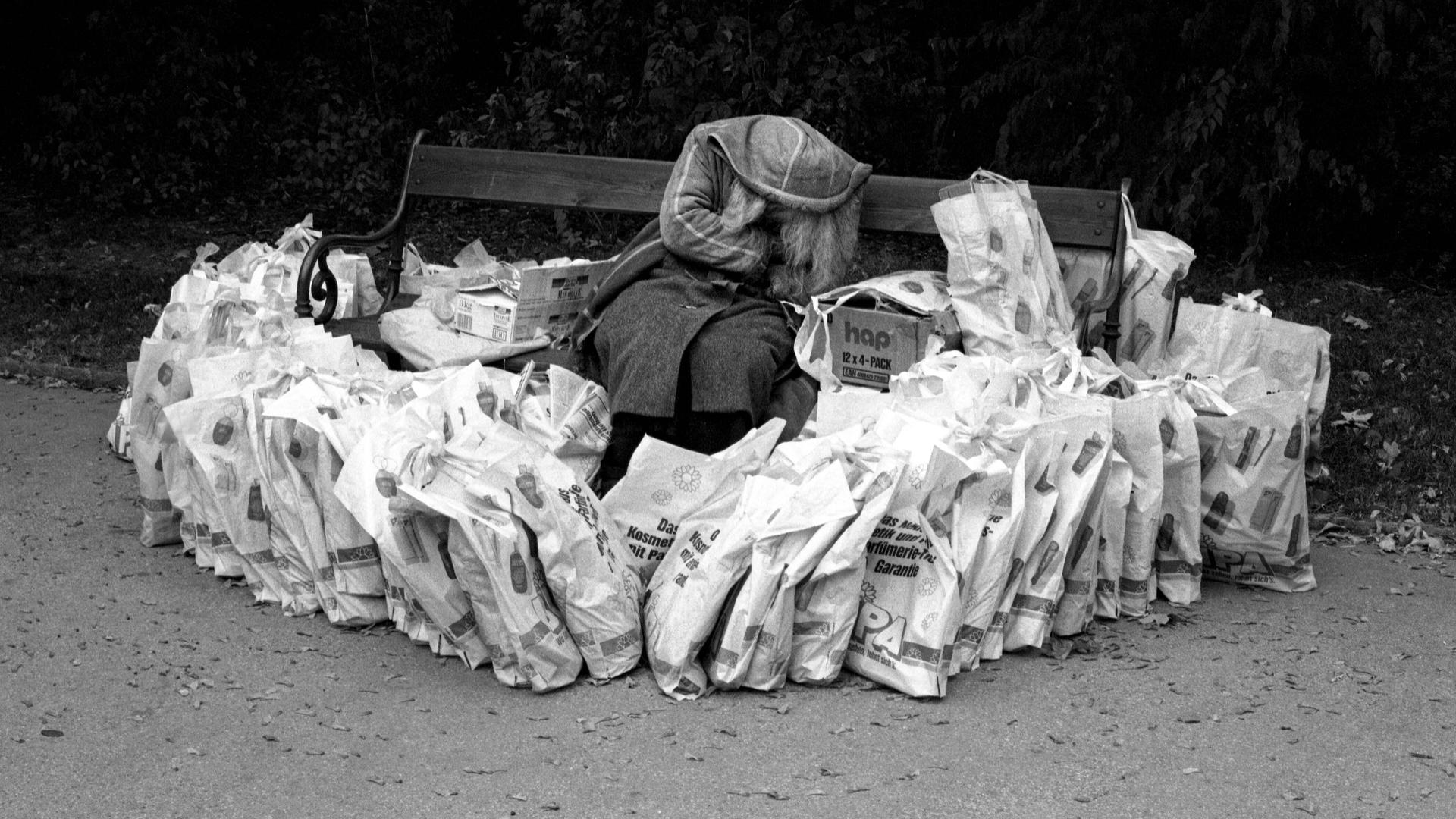 Eine obdachlose Frau versteckt sich auf einer Parkbank hinter einer Kapuze. Um sie herum liegen sehr viele Plastiktüten, aufgebaut wie eine Burg, 1989 .