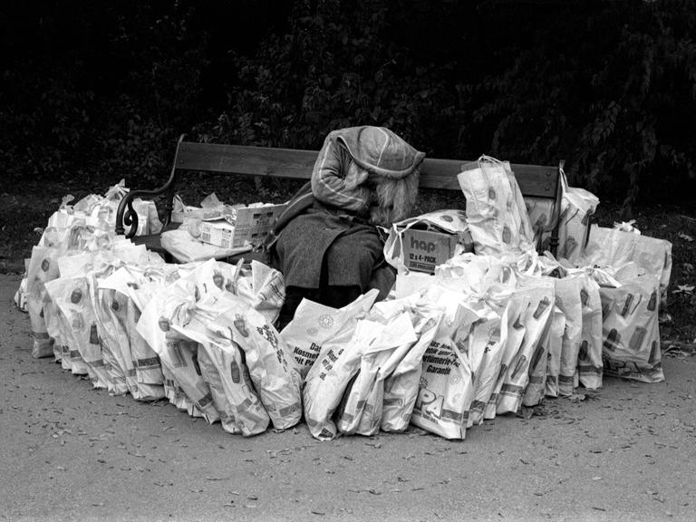 Eine obdachlose Frau versteckt sich auf einer Parkbank hinter einer Kapuze. Um sie herum liegen sehr viele Plastiktüten, aufgebaut wie eine Burg, 1989 .