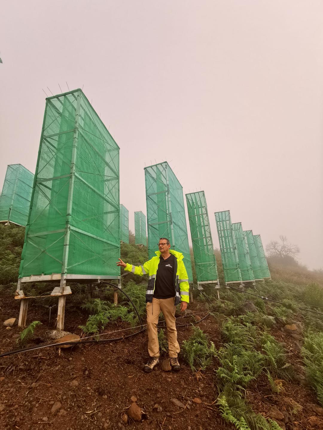 Mann in einer gelben Jacke steht neben hohen kastenförmigen Netzkonstruktionen: Biologe Paco González neben seinen Nebelfängern.