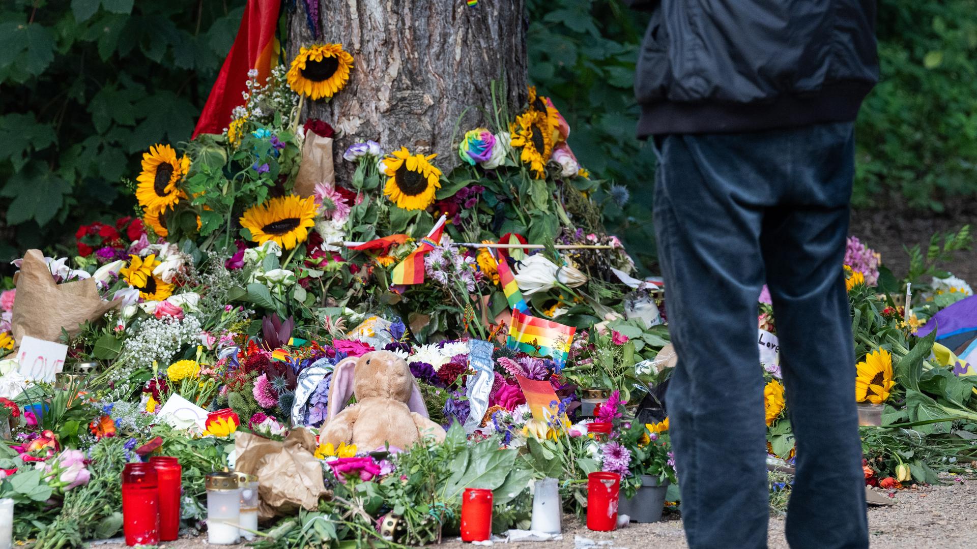 Trauernde stehen im Tiergarten an einem Baum, an dem unterschiedliche Gegenstände zum Gedenken an die Opfer des Anschlags auf den CSD abgelegt wurden.
