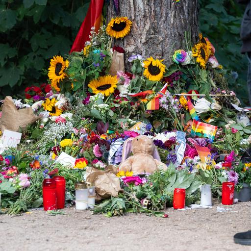 Trauernde stehen im Tiergarten an einem Baum, an dem unterschiedliche Gegenstände zum Gedenken an die Opfer des Anschlags auf den CSD abgelegt wurden.