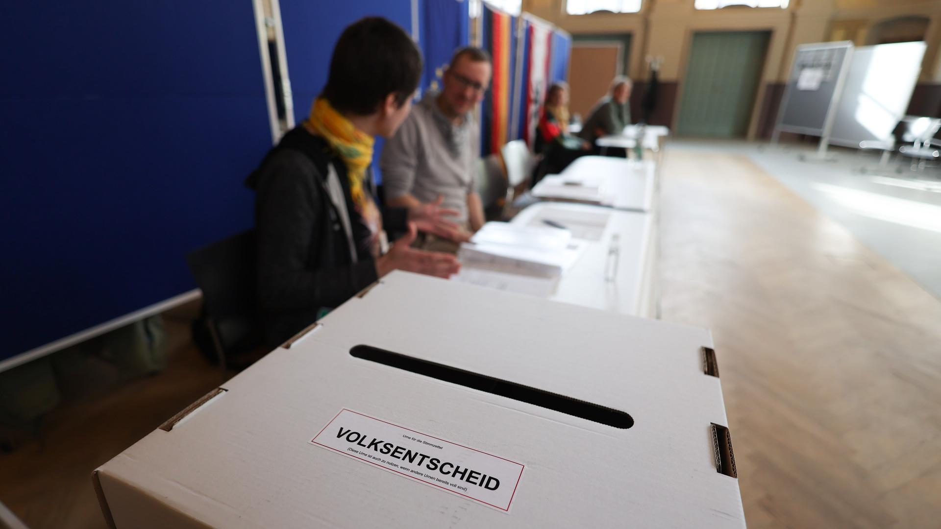 Das Foto zeigt eine Wahlurne. Auf dem Behälter ist ein Aufkleber mit der Aufschrift "Volksentscheid". Im Hintergund sind verschwommen Wahlhelfer zu sehen.
