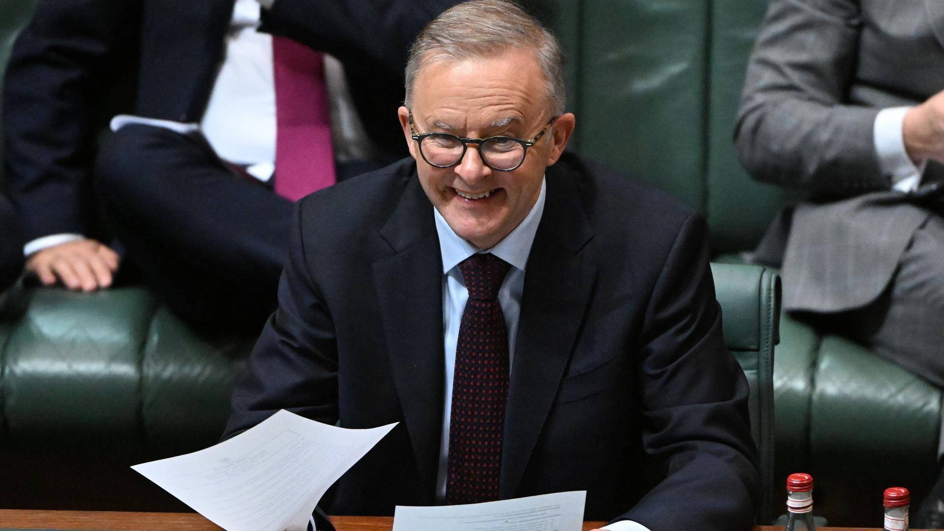 Der australische Premierminister Albanese sitzt an einem Tisch, hat zwei Blätter in der Hand und lächelt.