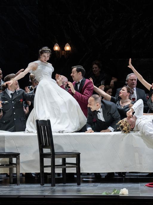 Auf einem langen Tisch steht eine Frau mit einem weißen Kleid umgeben von Feiernden.