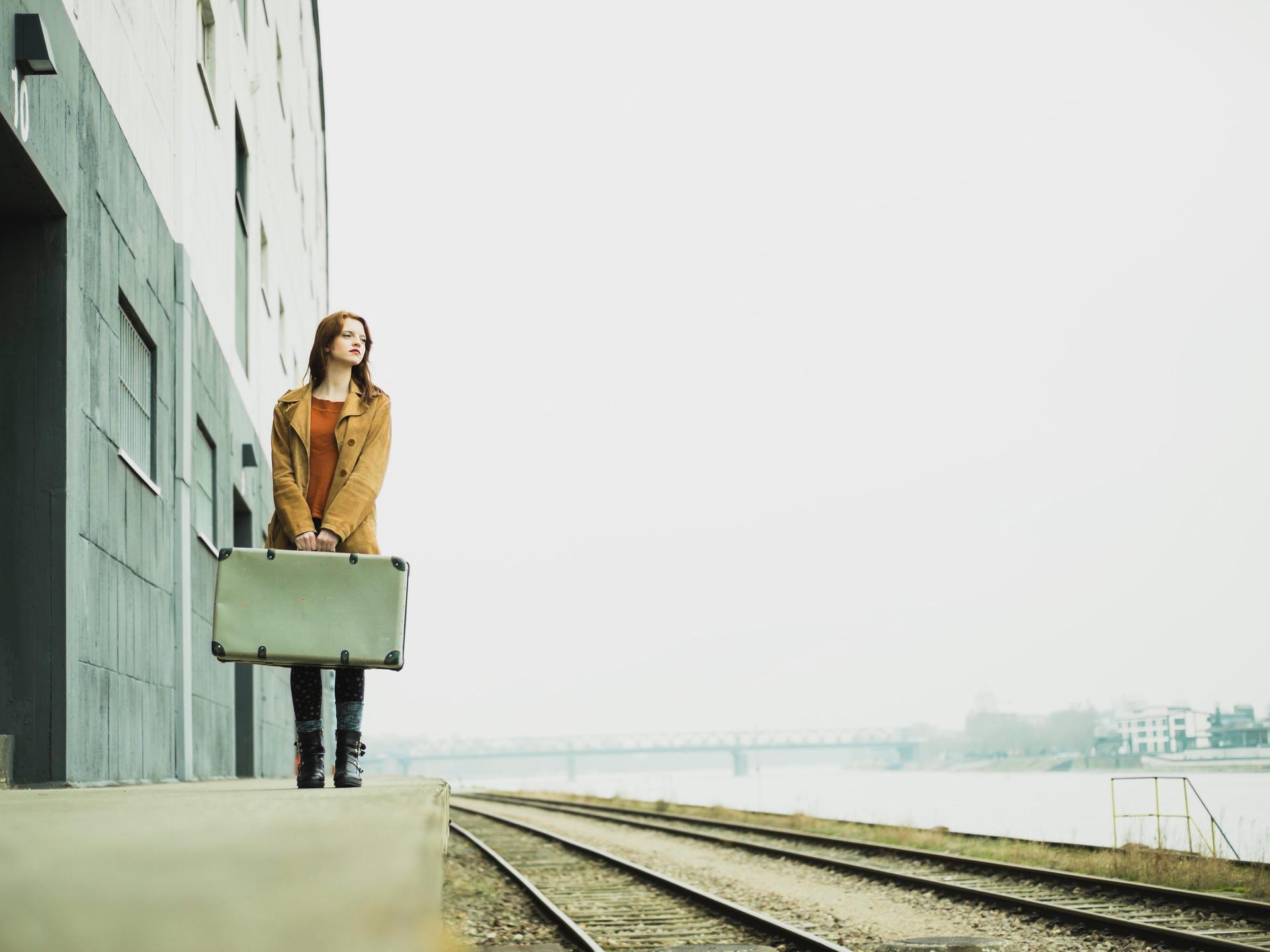 Eine Frau steht mit einem Koffer in der Hand an einem Bahngleis.
