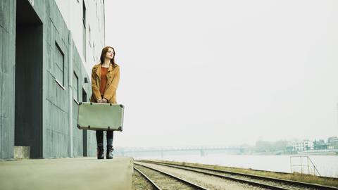 Eine Frau steht mit einem Koffer in der Hand an einem Bahngleis. Eine Frau steht mit einem Koffer in der Hand an einem Bahngleis.
