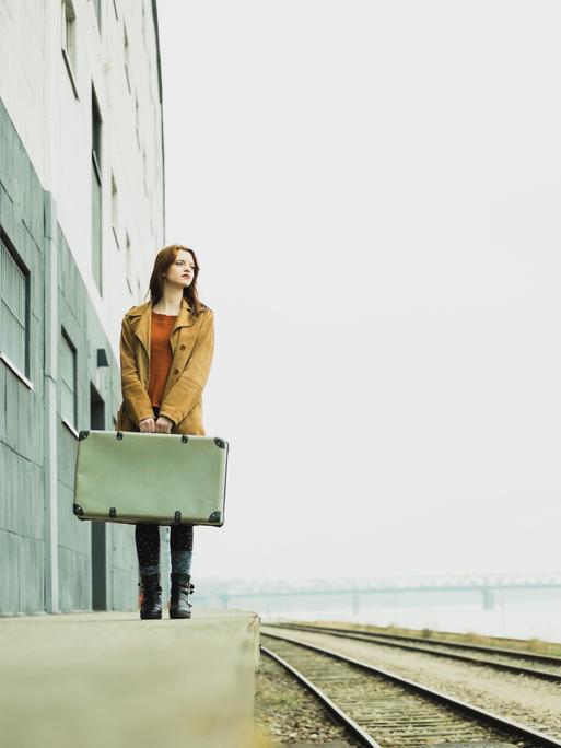 Eine Frau steht mit einem Koffer in der Hand an einem Bahngleis.