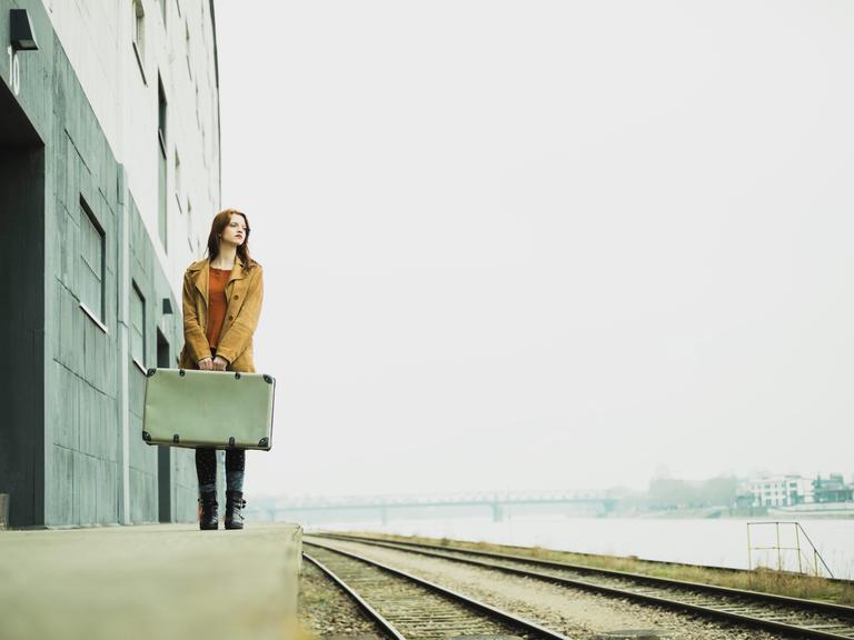Eine Frau steht mit einem Koffer in der Hand an einem Bahngleis.