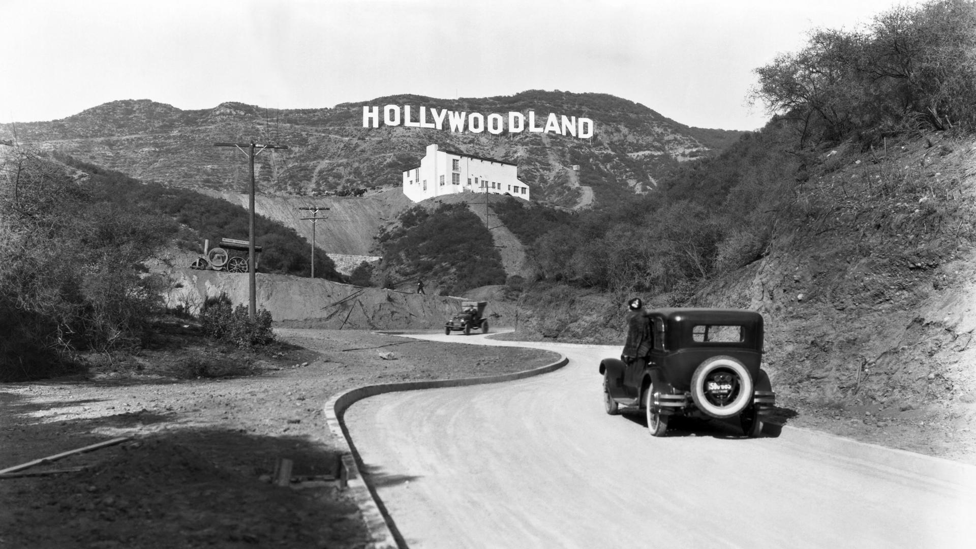 Ein Schild wirbt für die Eröffnung der Hollywoodland-Siedlung in den Hügeln am Mulholland Drive mit Blick auf Los Angeles um 1924. Darunter ein passierendes Auto und ein weißes Gebäude, die Kanst Art Gallery, die am 1. April 1924 eröffnet wurde.


