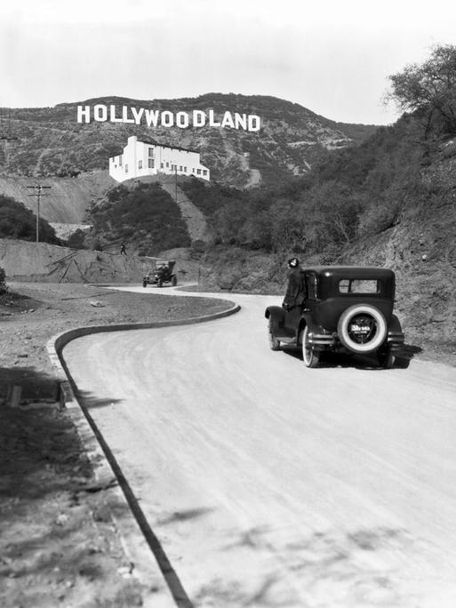 Ein Schild wirbt für die Eröffnung der Hollywoodland-Siedlung in den Hügeln am Mulholland Drive mit Blick auf Los Angeles um 1924. Darunter ein passierendes Auto und ein weißes Gebäude, die Kanst Art Gallery, die am 1. April 1924 eröffnet wurde.

