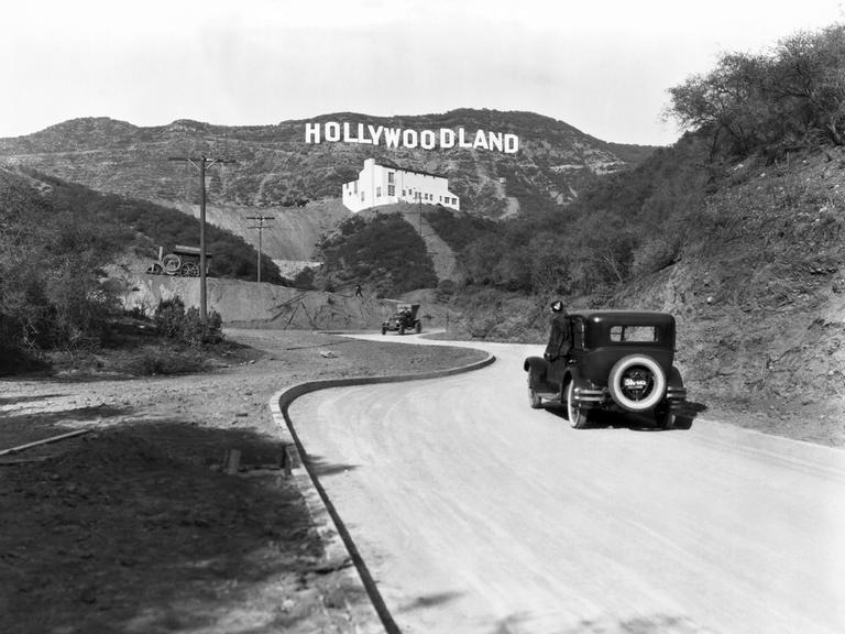 Ein Schild wirbt für die Eröffnung der Hollywoodland-Siedlung in den Hügeln am Mulholland Drive mit Blick auf Los Angeles um 1924. Darunter ein passierendes Auto und ein weißes Gebäude, die Kanst Art Gallery, die am 1. April 1924 eröffnet wurde.

