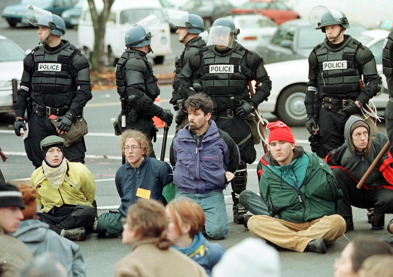 Auf der Straße sitzen junge Demonstrant*innen. Dahinter stehen Polizisten mit Schutzhelmen.