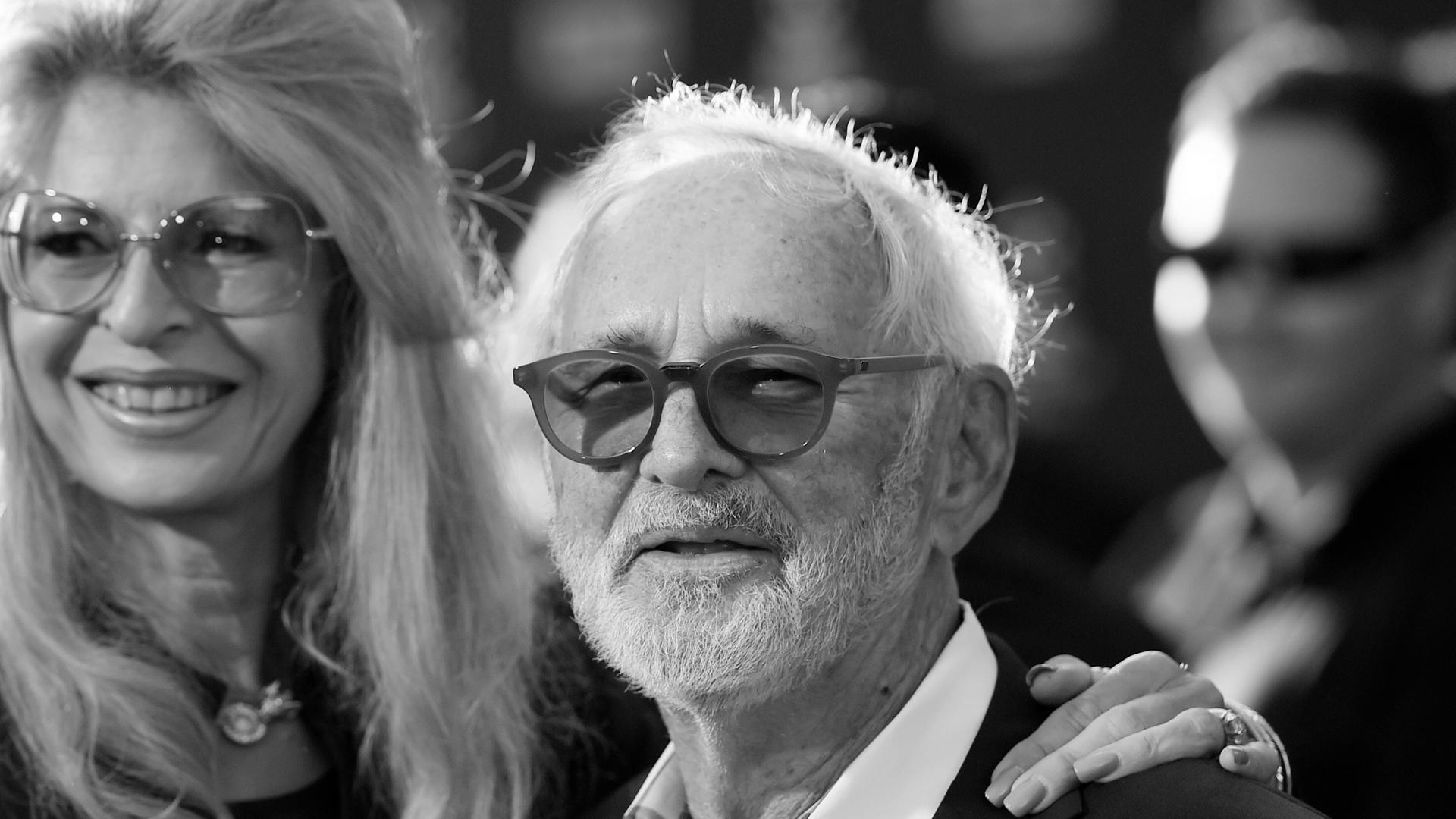 Norman Jewison mit weißem schütteren Haar, weißem Bart und dunkler Brille. Seine Frau legt einen Arm auf seine Schulter.