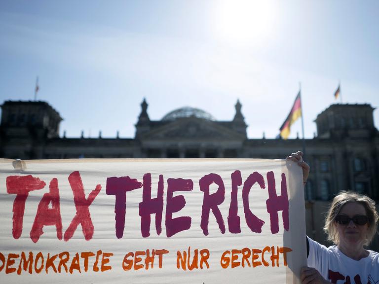 Eine Frau hält ein Transparent mit der Aufschrift "Tax the rich - Demokratie geht nur gerecht" vor dem Deutschen Bundestag hoch