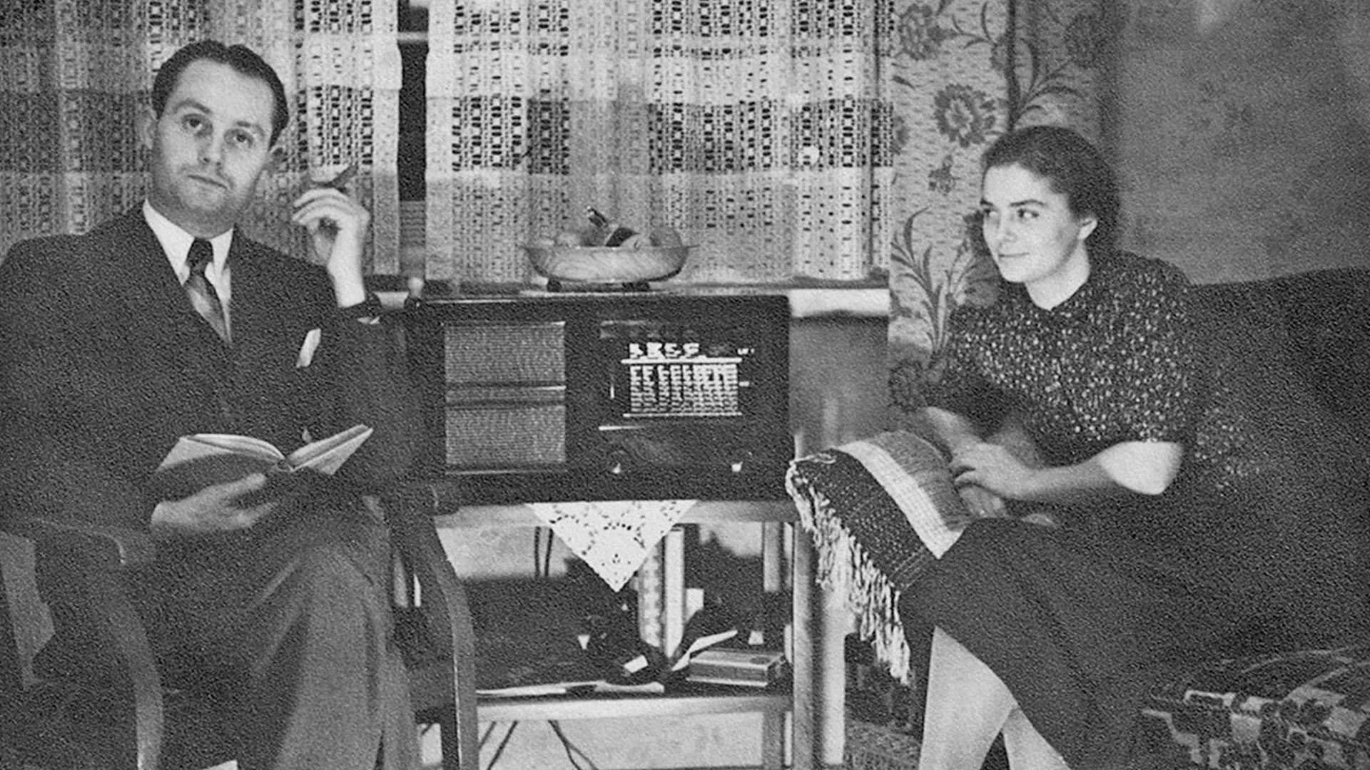 Ein gut bürgerliches Ehepaar sitzt in ihrer Wohnung an einem Volksempfänger.