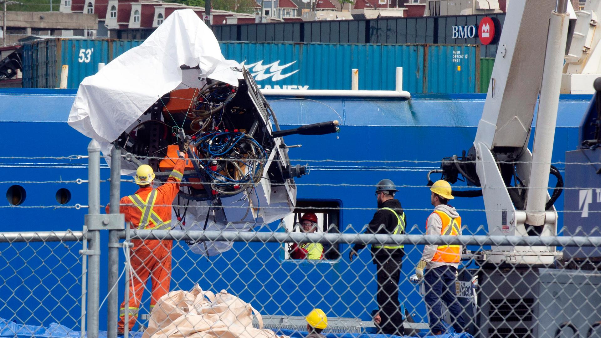 Im Vordergrund ein Zaun in einem Hafen, dahinter stehen Arbeiter vor einem Schiff. Einer hält ein per Kran entladenes Wrackteil fest.