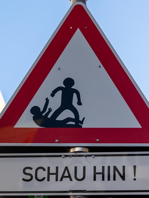 Dreieckiges Warnschild mit rotem Rand und weißem Hintergrund. Im Inneren des Dreiecks befinden sich schwarze Silhouetten von zwei Figuren, von denen eine aggressiv erscheint und die andere defensiv reagiert.