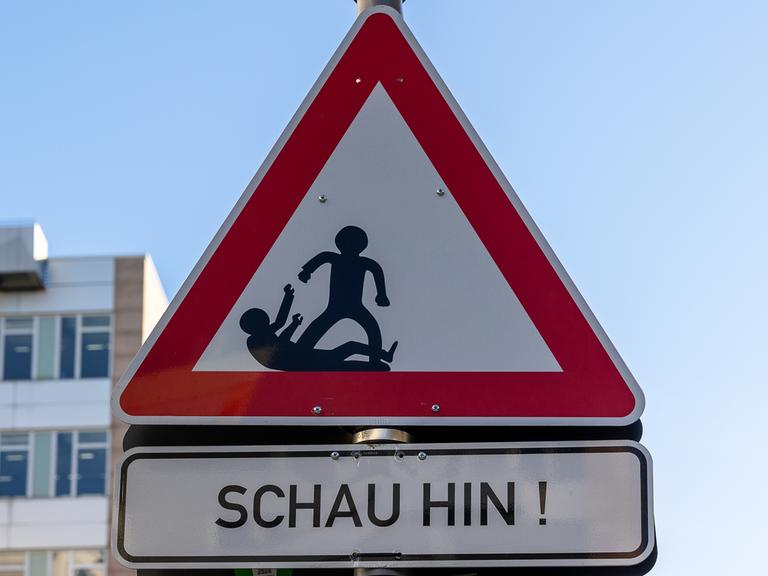 Dreieckiges Warnschild mit rotem Rand und weißem Hintergrund. Im Inneren des Dreiecks befinden sich schwarze Silhouetten von zwei Figuren, von denen eine aggressiv erscheint und die andere defensiv reagiert.
