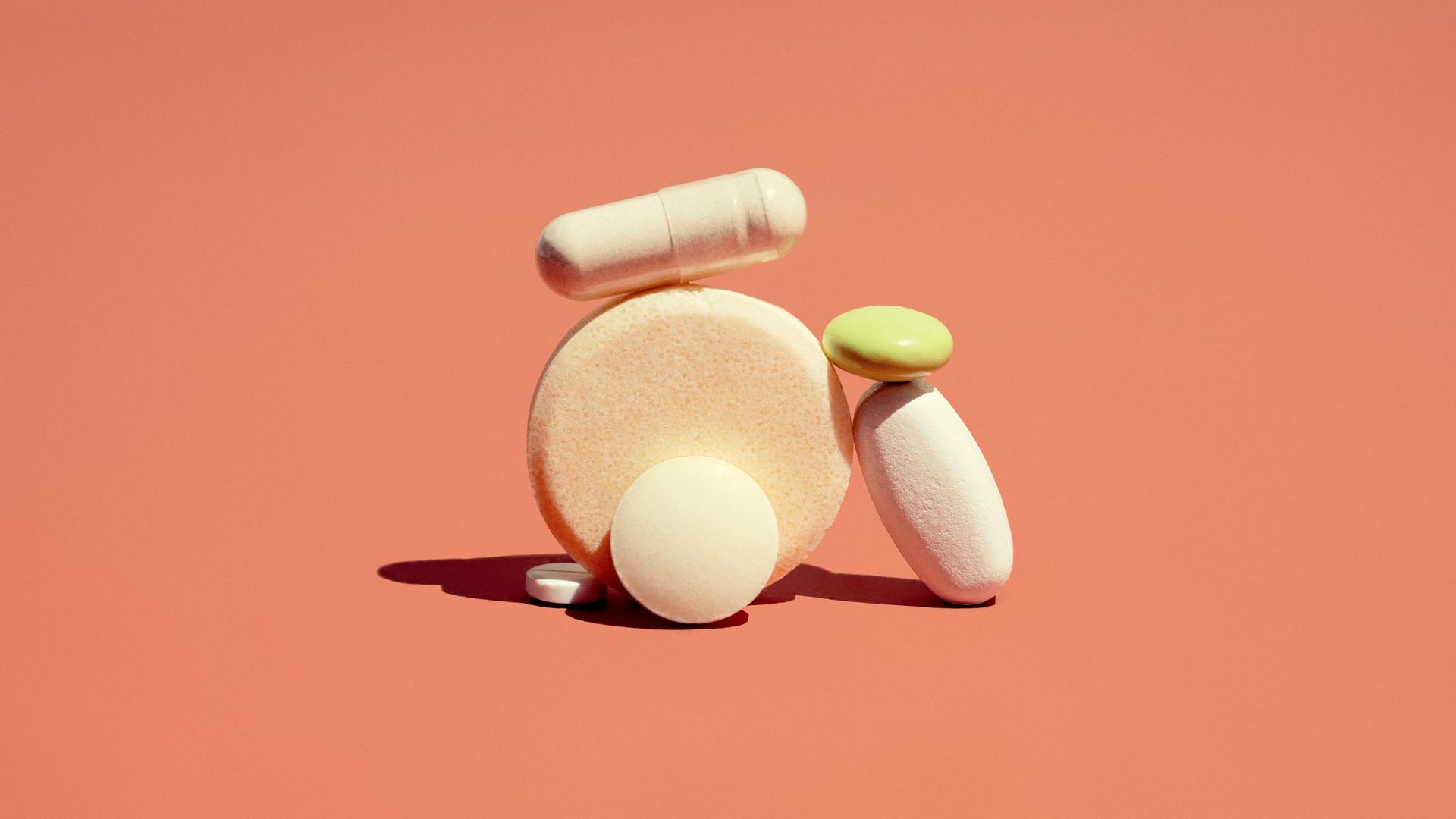 Medikamente in Form von Pillen und Zäpfchen liegen zu einem Türmchen gestapelt auf einem orange farbigem Hintergrund. 