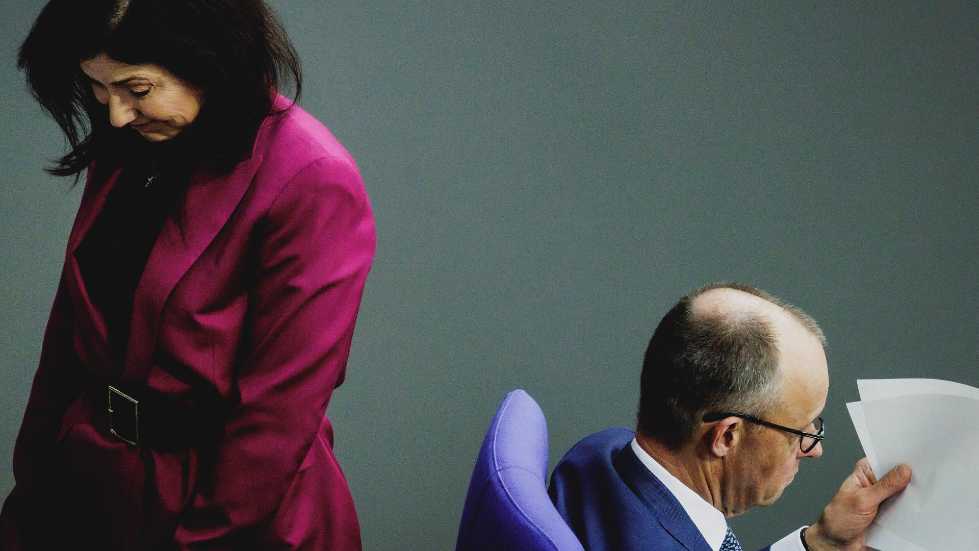 Bundeskanzler Merz (r.) und Bundeswirtschaftsministerin Reiche drehen einander den Rücken zu. Reiche steht und trägt ein magentafarbenes Jackett. Merz sitzt und schaut in ein Dokument. 