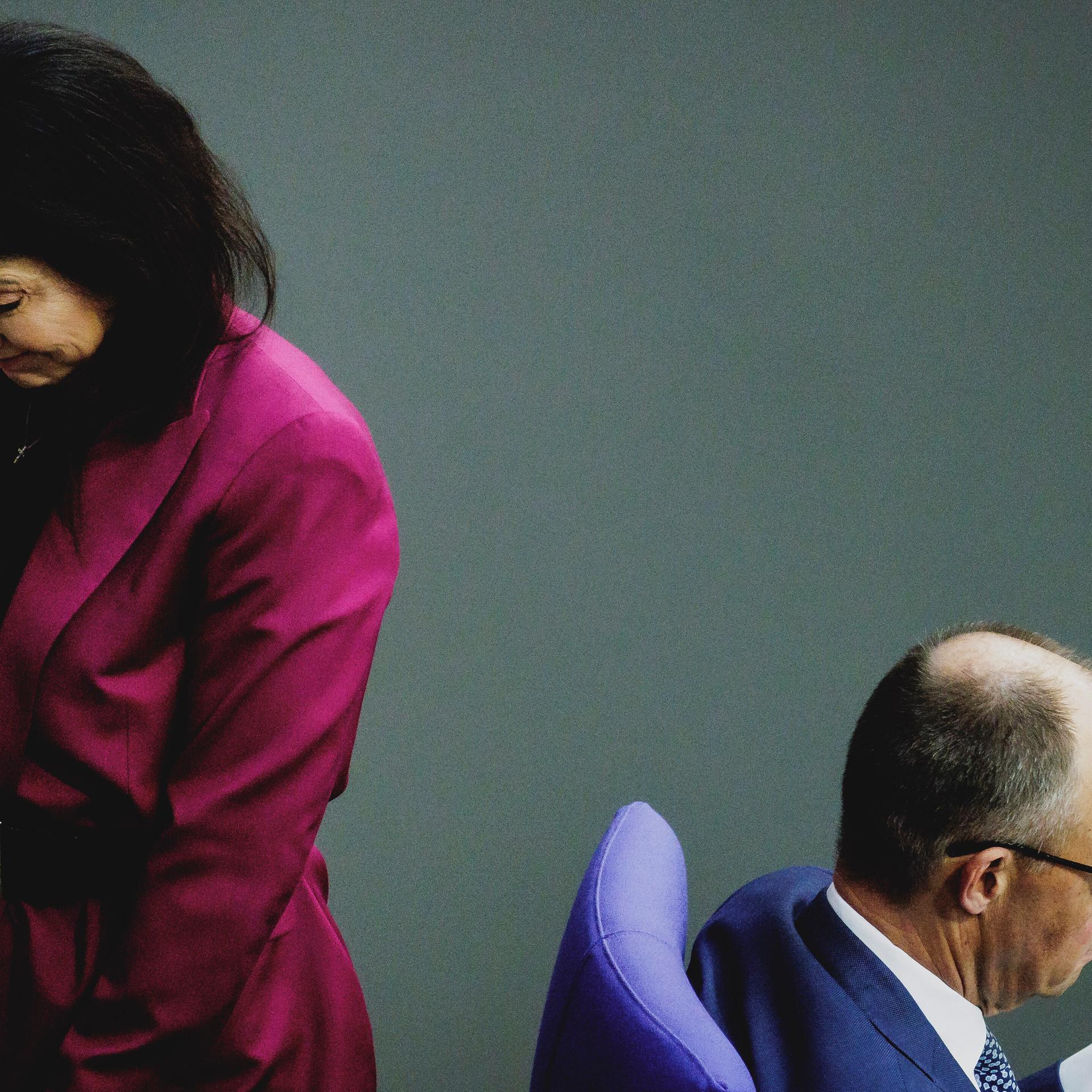 Die beiden drehen einander den Rücken zu. Reiche steht und trägt ein magentafarbenes Jackett. Merz sitzt und schaut in eine Dokument. 