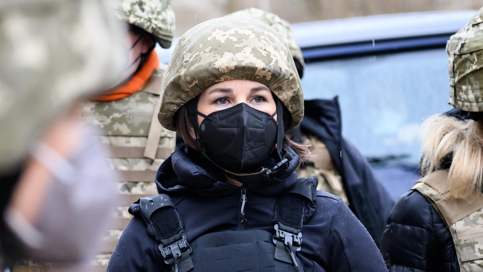 Außenministerin Annalena Baerbock im Konfliktgebiet Donbass in der Ukraine. Sie trägt eine schwarze FFP2-Maske, einen Tarnhelm und eine schwarze Schutzweste. Sie ist umringt von Menschen, die ebenfalls Tarn- und Schutzkleidung tragen.