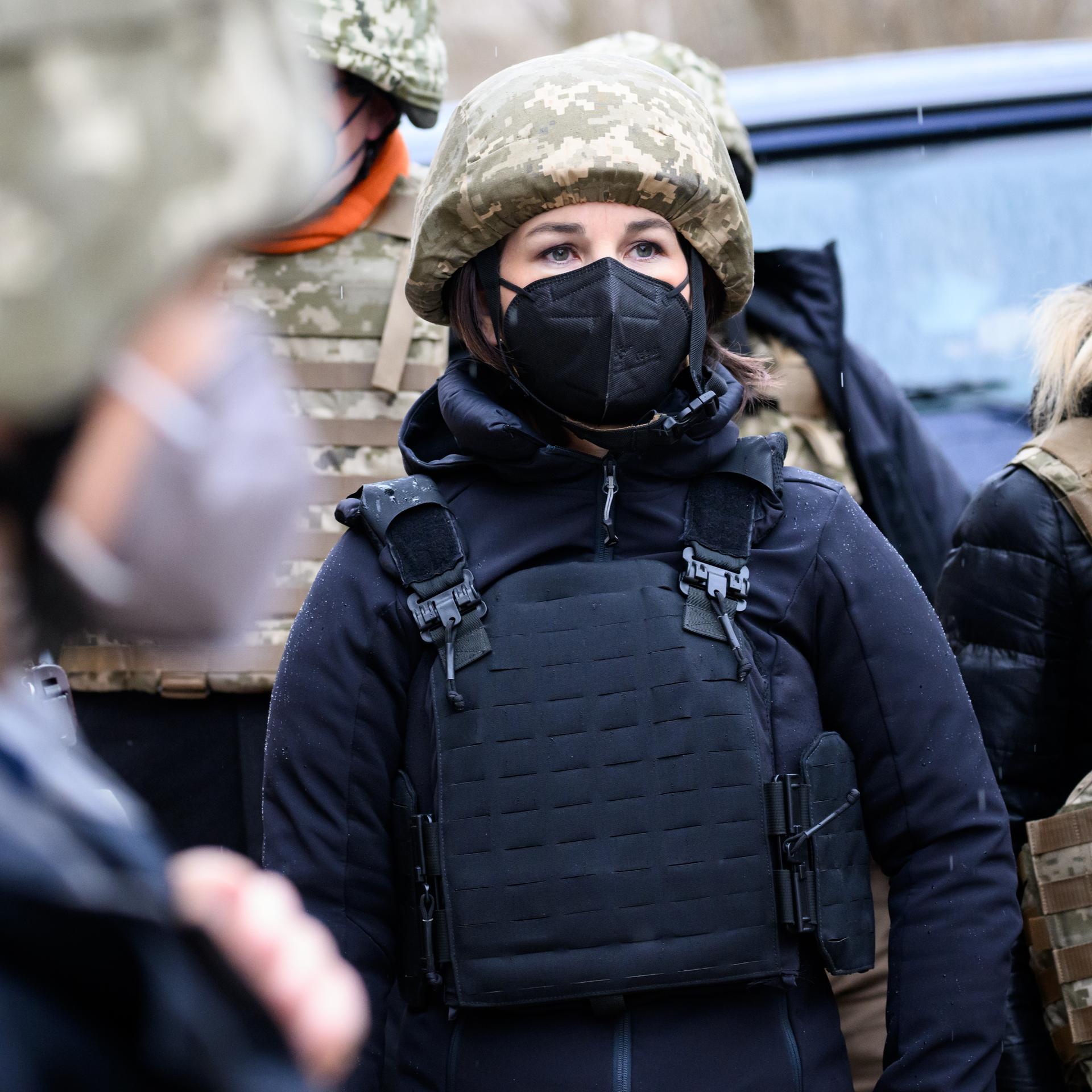 Außenministerin Annalena Baerbock im Konfliktgebiet Donbass in der Ukraine. Sie trägt eine schwarze FFP2-Maske, einen Tarnhelm und eine schwarze Schutzweste. Sie ist umringt von Menschen, die ebenfalls Tarn- und Schutzkleidung tragen.