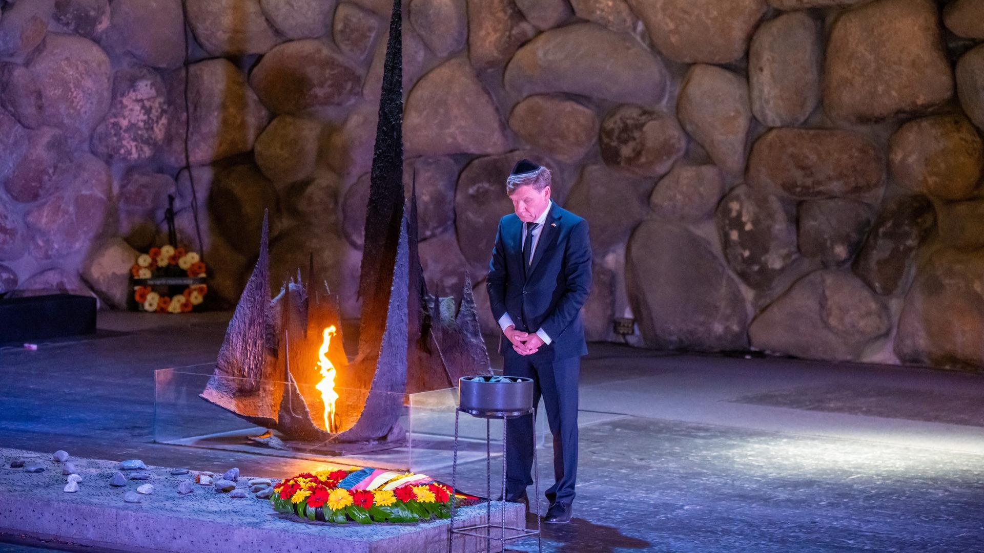 Ein Mann in Anzug und mit Kippa steht mit gefalteten Händen in einer steinernen Halle. Er blickt nachdenklich auf einen am Boden liegenden Blumenkranz. Im Hintergrund brennt eine Flamme. Ein Mann in Anzug und mit Kippa steht mit gefalteten Händen in einer steinernen Halle. Er blickt nachdenklich auf einen am Boden liegenden Blumenkranz. Im Hintergrund brennt eine Flamme.