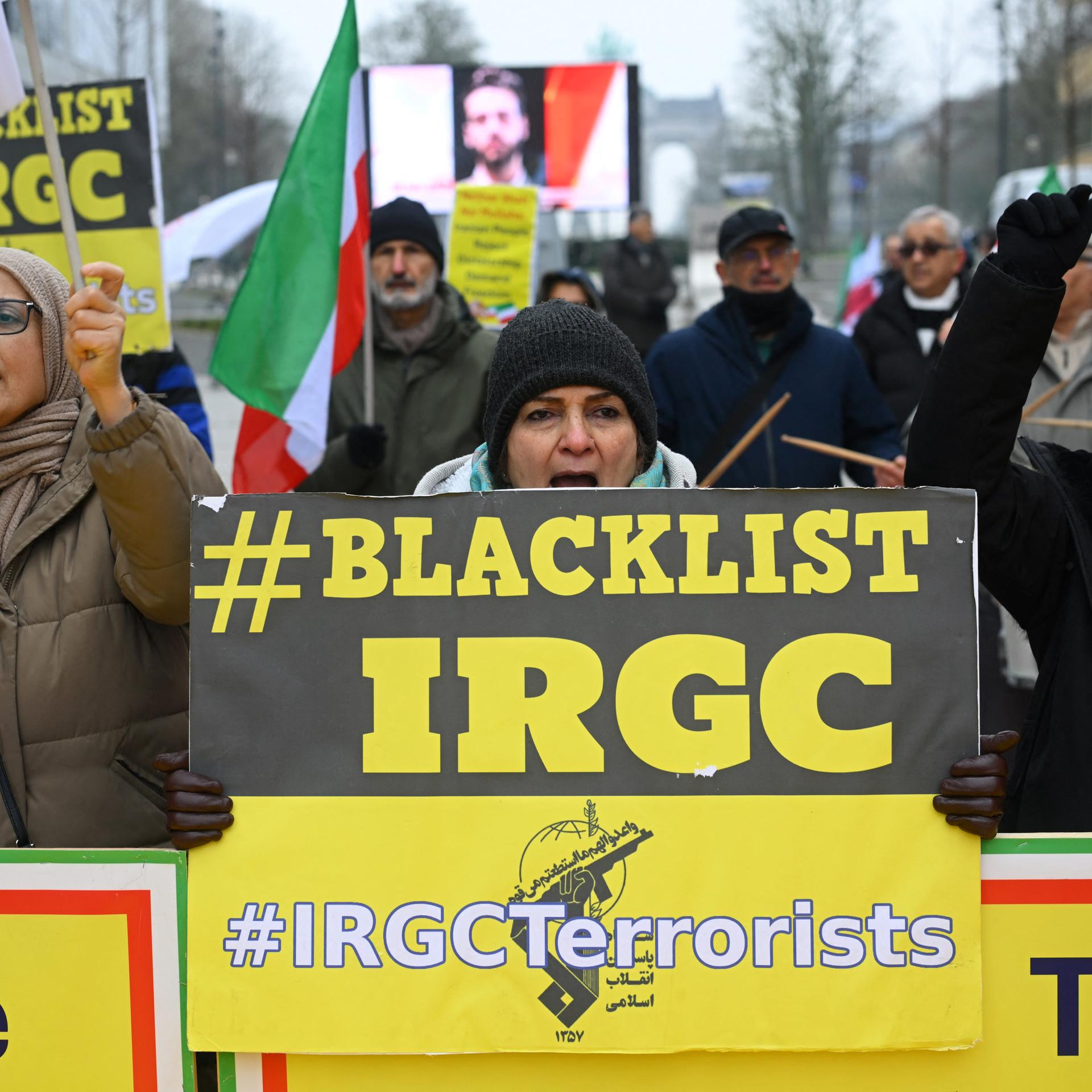 Das Bild zeigt demonstrierende Menschen auf einer Straße in Brüssel. Sie halten gelbe Schilder mit dem Aufdruck "#BLACKLIST IRGC" und fordern damit die Einstufung der iranischen Revolutionsgarde als Terrororganisation.